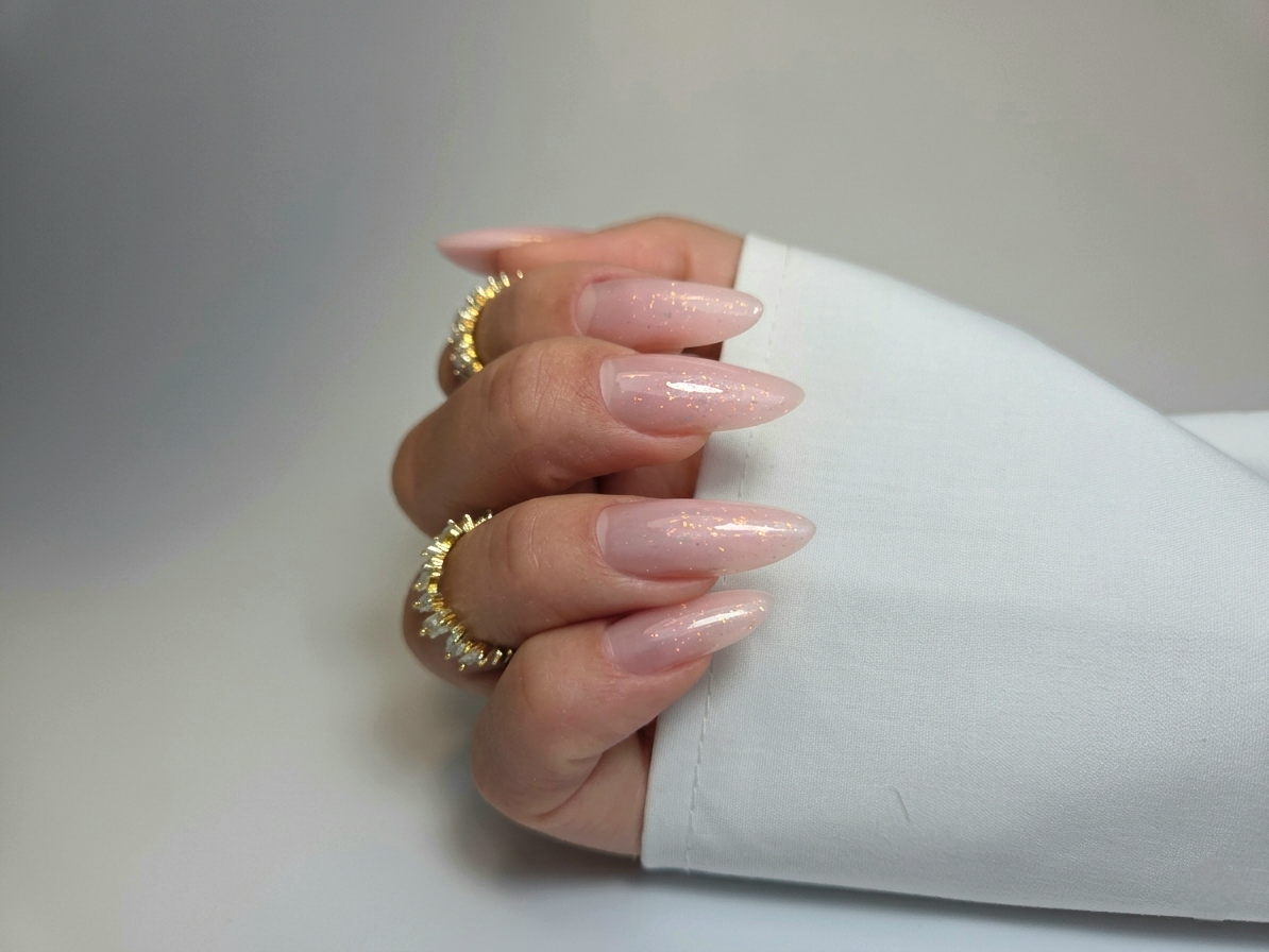 Lange amandelvormige nagels in zacht roze met glitter, met gouden ringen